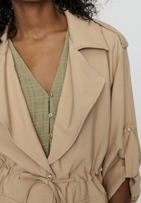 Kvinna iförd en beige trenchcoat knuten i midjan över en olivgrön blus med knappar och ett subtilt rutmönster.