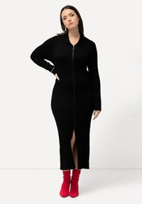 Robe noire en maille côtelée avec fermeture éclair à l'avant, col et fente latérale. Associée à des bottes de cheville rouges à la coupe décontractée.