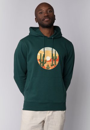 Mann trägt dunkelgrünen Hoodie mit Grafik eines Wüstensonneruntergangs und Kaktus, kombiniert mit hellbeigen Hosen, steht vor neutralem Hintergrund.