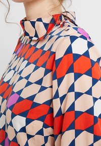 Blouse à motifs géométriques avec un col montant, en rouge, bleu et blanc. Confectionnée dans un tissu léger et lisse avec une coupe décontractée.
