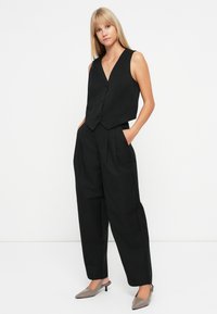 Gilet senza maniche nero con scollo a V e bottoni, abbinato a pantaloni neri a vita alta con pieghe. Scarpe grigie con tacco a kitten.