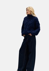 Gruby granatowy sweter z dzianiny o wysokim kołnierzu, z teksturowanym wzorem warkocza. W połączeniu z szerokimi, ciemnoniebieskimi dżinsami.