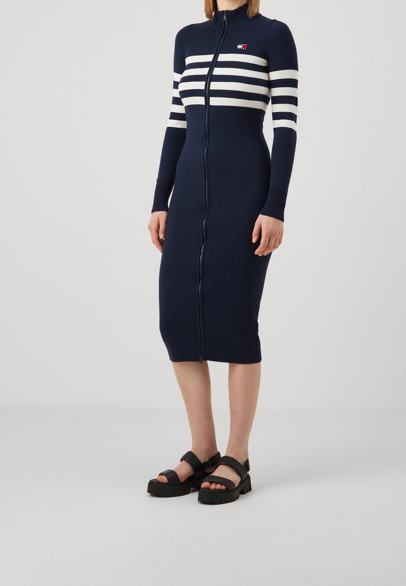 Femme portant une robe midi bleu marine à manches longues avec fermeture éclair, rayures blanches horizontales sur la poitrine, et des sandales noires à larges brides épaisses.