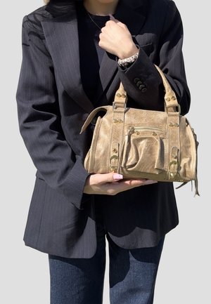 Donna con blazer scuro e jeans che tiene una piccola borsa in camoscio color cuoio con dettagli in metallo dorato e tasca anteriore.