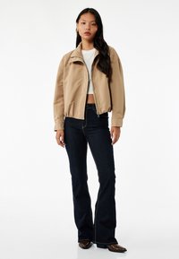 Beżowa kurtka bomberka z zamkiem błyskawicznym, ściągaczem u dołu i przy kołnierzu, noszona na białej bluzce i ciemnych dzwonach. Gładka tekstura tkaniny.