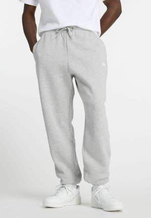 Person iført lysegrå joggingbukser med snøre i taljen, parret med hvide sneakers og en hvid T-shirt, stående med hænderne i lommerne.