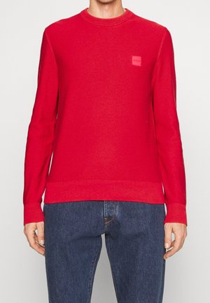 Roter Strickpullover mit langen Ärmeln, Rundhalsausschnitt, strukturiertem Stoff und einem kleinen geprägten Logo-Label auf der Brust. Kombiniert mit dunklen Jeans.