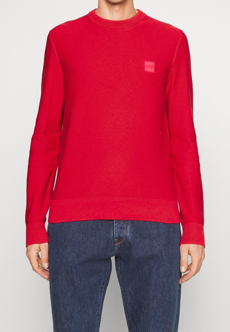 Jersey de punto rojo con mangas largas, escote redondo, tejido texturizado y un pequeño parche de logo en relieve en el pecho. Combinado con pantalones vaqueros oscuros.