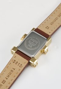 Gold-tone rectangular watch with a silver metallic backing, brown leather strap, and engraved details on the case and strap underside.