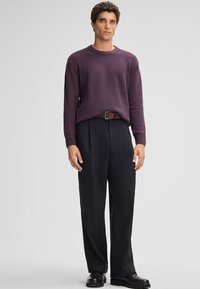 Maglione viola lavorato a maglia con finitura testurizzata, abbinato a pantaloni neri con vestibilità ampia e scarpe in pelle nera. La cintura marrone conferisce un contrasto.