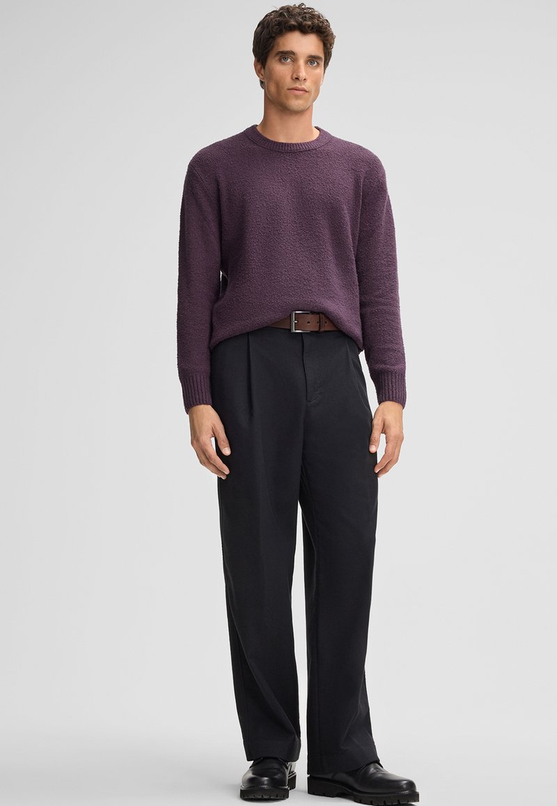 Maglione viola lavorato a maglia con finitura testurizzata, abbinato a pantaloni neri con vestibilità ampia e scarpe in pelle nera. La cintura marrone conferisce un contrasto.