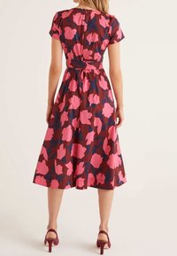 Robe brune avec un motif floral rose et des accents bleus, manches courtes, taille cintrée et jupe fluide. Agrémentée de chaussures à talons hauts.