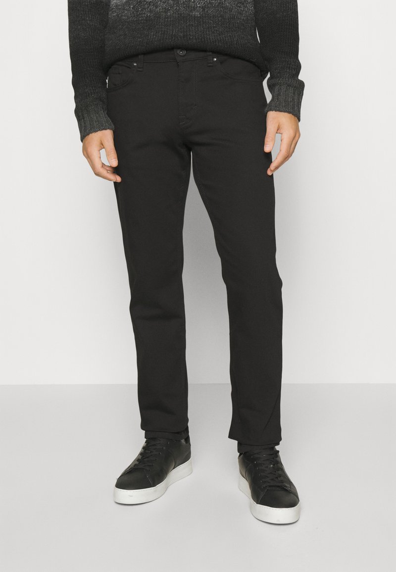 Musta slim-fit farkut, joissa on sileä pinta, viisi taskua ja nappikiinnitys, yhdistettynä mustiin tennareihin, joissa on valkoiset pohjat.
