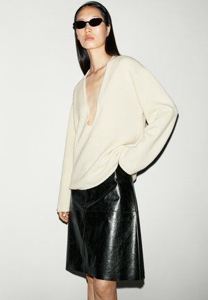 Donna che indossa occhiali da sole neri, maglione crema con scollo a V profondo e gonna di pelle nera su sfondo chiaro uniforme.