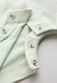 Gros plan de tissu vert clair avec des boutons-pression métalliques argentés sur des bords qui se chevauchent, montrant les coutures et la texture.