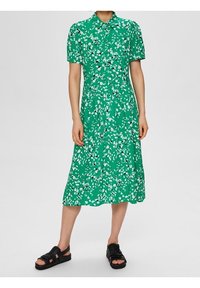 Vestito midi verde con maniche corte, caratterizzato da un motivo floreale in bianco e nero, scollatura abbottonata e gonna svasata. In uso sandali neri.