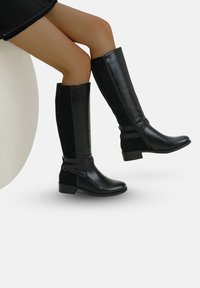Stivaletti neri al ginocchio con tomaia in morbida pelle sintetica, chiusura con zip laterale e dettagli decorativi con cinturini. Tacco basso a blocco.