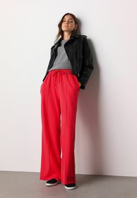 Rote weite Hosen mit einem Bindegürtel, kombiniert mit einem grauen Pullover und einer schwarzen Lederjacke; schwarze Sneakers runden das Outfit ab.