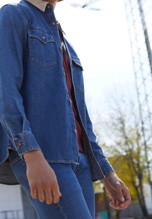 Person, die eine blaue Jeansjacke über einem roten Hemd und blauen Jeans trägt, steht im Freien mit Bäumen und einem Turm im Hintergrund.