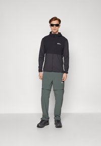 Svart og grå zip-up jakke med hette, parret med lysegrønne bukser med glidelåser. Modellen har på svarte sko og solbriller.