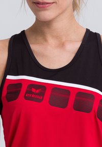 Top de tirantes rojo y negro con escote redondo, rayas blancas de acento y detalles gráficos texturizados, con el logo de "erima" exhibido de forma prominente.