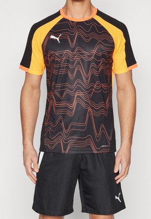 Mand iført sort og orange Puma sportstrøje med røde bølgemønstre og sorte Puma shorts, stående med afslappede arme.