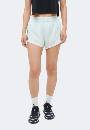 Žena oblečená v svetlosivých športových šortkách s čiernym logom Nike, biele rebrované ponožky a čierne bežecké topánky, stojaca pred jednoduchým pozadím.