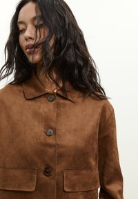 Žena s dlhými tmavými vlasmi, oblečená v hnedej semišovej bunde zapínanej na gombíky s klopovými vreckami a korálkovým náhrdelníkom, mierne naklonená hlavu smerom nadol.