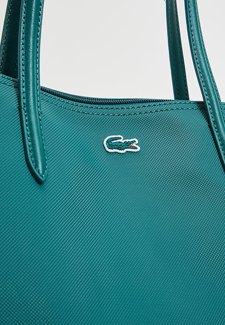 Bolso de mano texturizado en color azul verdoso con asas lisas, que presenta un pequeño logo de cocodrilo bordado en blanco. Diseño estructurado con cierre de cremallera.