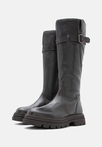 Ruske nahke čizme do kolena s teksturiranim finišem, trakom i kopčom, te debelim gumiranim đonom s izraženim uzorkom.