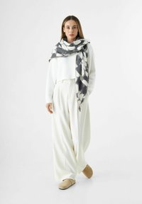 Femme portant un pantalon large blanc, un pull blanc, des chaussures beiges et un grand foulard noir et blanc à motifs drapé sur les épaules.