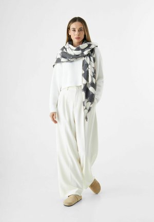 Vrouw gekleed in witte wijde broek, witte trui, beige schoenen en een grote zwart-wit gestreepte sjaal over de schouders gedrapeerd.