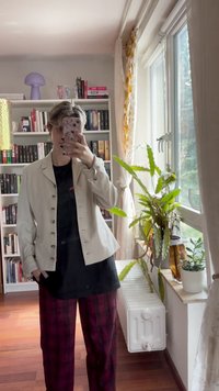 Light beige jacket with metal buttons, worn over a black t-shirt, paired with dark red plaid pants. Indoor setting with plants and bookshelf.