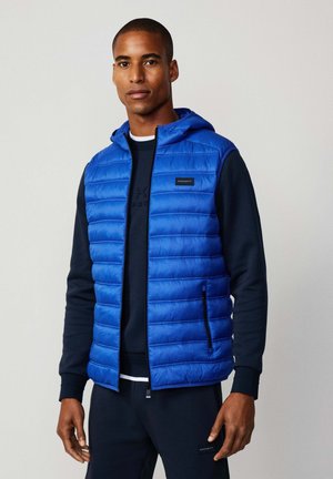 Mann mit blauer gesteppter ärmelloser Weste über navy Sweatshirt und Hose, der vor einem hellen, einfarbigen Hintergrund steht.
