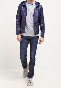 Muž kráčející v námořnickém kapucovém kabátu přes světle šedé polo triko, tmavě modrých džínách a světle šedých teniskách před jednobarevným pozadím.