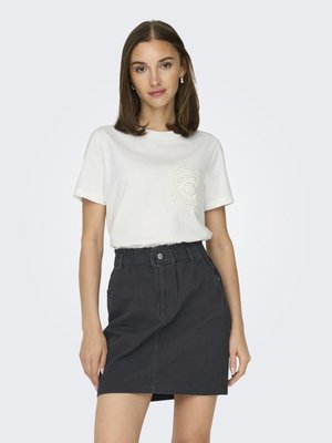 Vrouw draagt een wit shirt met korte mouwen en een kanten zak, samen met een zwarte hoge taille rok, staand tegen een effen lichte achtergrond.