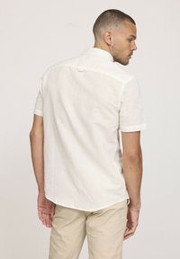 Un homme portant une chemise blanche à col, à manches courtes, et un pantalon beige, tourné légèrement sur le côté, montrant le dos et le profil.