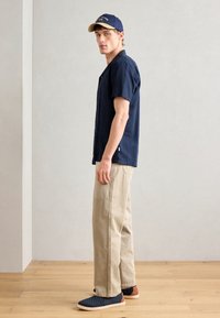 Camicia blu navy a maniche corte, pantaloni cargo beige chiaro, sneakers blu navy con dettagli marroni e un cappellino blu navy con visiera beige. Design minimalista.