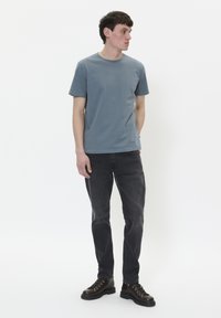 Camiseta de algodón gris, de cuello redondo, combinada con jeans gris oscuro y botas negras con cordones. El modelo está de pie sobre un fondo blanco liso.
