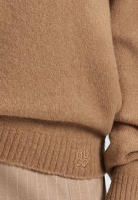 Pull beige de la marque HUGO avec une texture douce, des poignets et un ourlet côtelés, avec un logo brodé en bas à gauche.