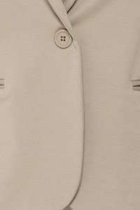 Béžové textúrované sako s jedným okrúhlym béžovým gombíkom zapnutým blízko vrchu, ukazujúce časť goliera a vreciek.