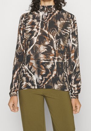 Femme portant un chemisier à manches longues avec un motif abstrait de feuilles marron, noir et blanc, associé à un pantalon vert olive.