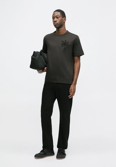 Tricou negru cu mânecă scurtă, având un design floral brodat pe piept, asortat cu pantaloni negri și adidași negri, purtând o geantă neagră.