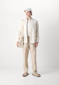Lettbeige knappert skjorte over en hvit topp; løse beige bukser, brune sandaler og en matchende cap; bærer en beige veske.