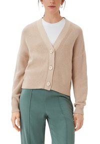 Beige Strickjacke mit V-Ausschnitt, ausgestattet mit drei Knöpfen und geripptem Muster. Kombiniert über einem weißen Oberteil und mit grünen Hosen.