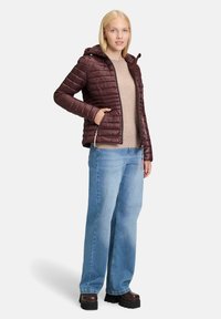 Burgundy pufferjakke med hætte, lynlåslukning og horisontale quiltede mønster, parret med en beige sweater og lyseblå wide-leg jeans.