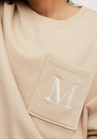 Beige Sweatshirt mit rundem Halsausschnitt, ausgestattet mit einer Fronttasche, die mit einem weißen gestickten Buchstaben "M" verziert ist. Weiche Textur, lässiges Design.