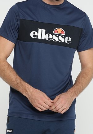 Mężczyzna w granatowej koszulce ellesse i pasujących szortach, ręce złożone na talii, widoczny na jasnym tle.