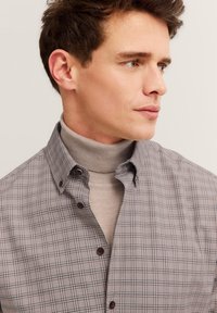Mann mit kurzen braunen Haaren, der einen beigen Rollkragenpullover unter einem beigen karierten Hemd trägt und nach rechts schaut, vor einem hellen Hintergrund.