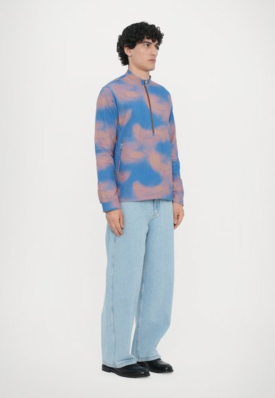 Zip-up, long-sleeve jacket featuring a blue and pink abstract pattern, ribbed texture, high collar, and zippered pocket detail. Paired with light jeans.